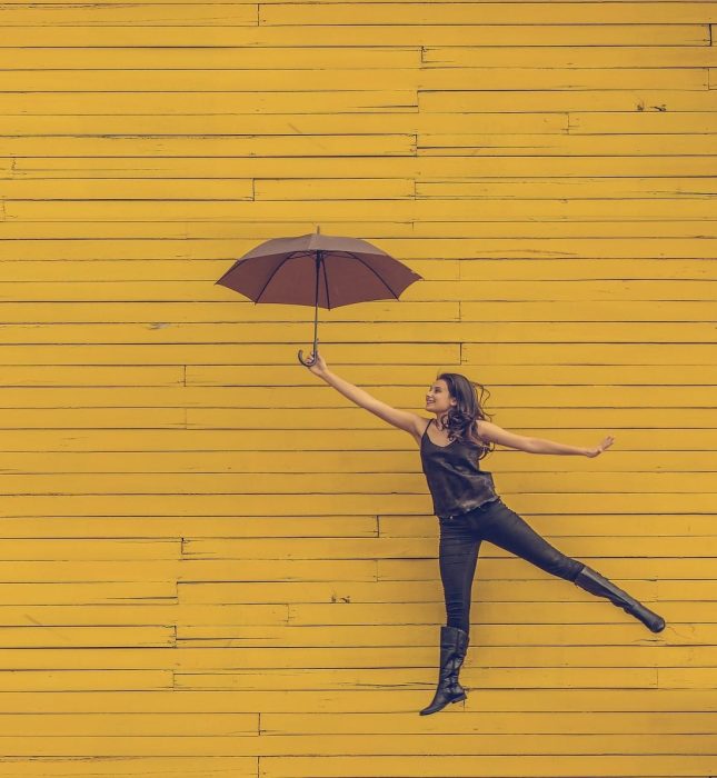 Joyful woman balancing on one leg with a maroon umbrella against a vibrant yellow wall.