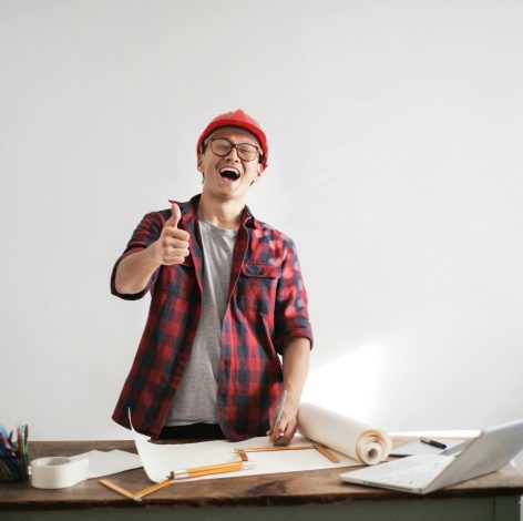 Young man in creative workspace smiling, giving thumbs-up, surrounded by design materials and blueprints.