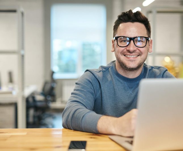 Young professional smiling at laptop in bright, modern office setting.
