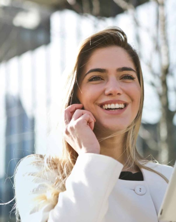 Cheerful young woman in white coat engaging in conversation against urban glass buildings.