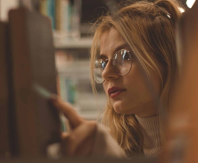 Thoughtful woman in cozy bookstore, reaching for a book, exuding warmth and curiosity.