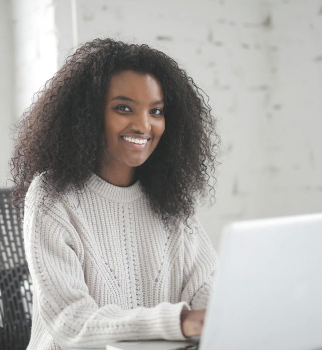 Young woman smiles while working on laptop in bright, cozy minimalist workspace.